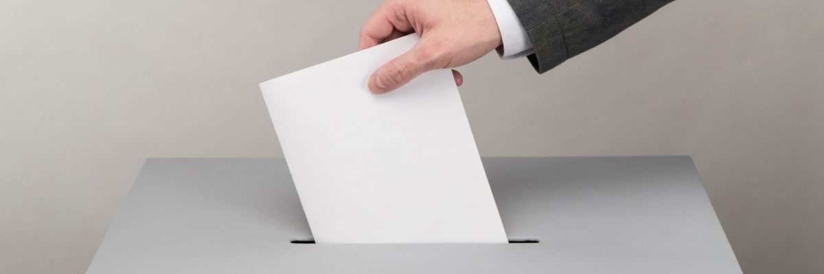 Dieses Bild zeigt eine Hand die ein Wahlkuvert in die Urne wirft.